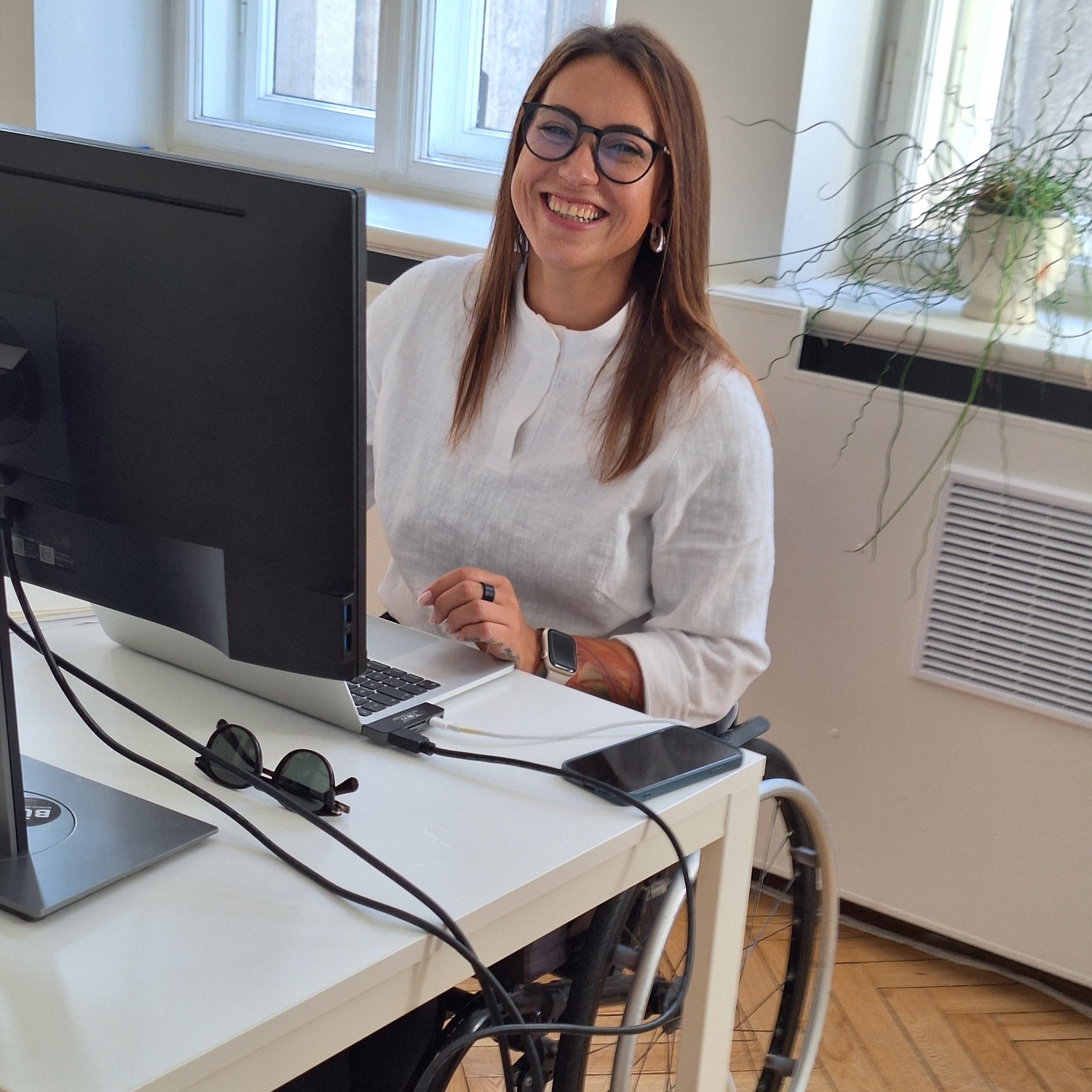 A smiling woman in a wheelchair at the desk in an office behind a computer. She is wearing glasses, smiling and ready to answer questions about adaptive clothing